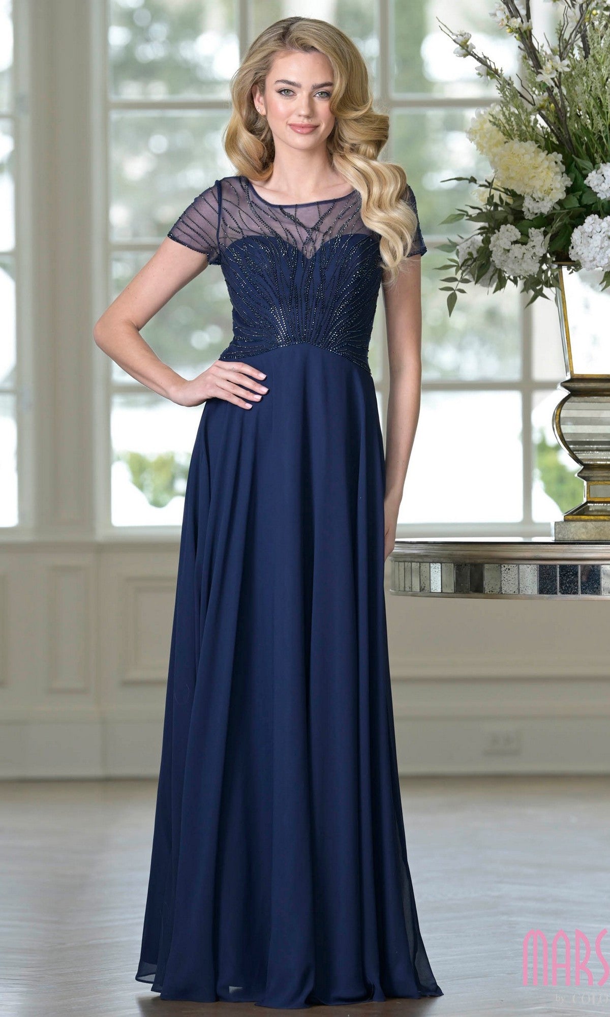 A woman stands indoors wearing the Marsoni M351 long formal dress in navy, featuring a chiffon A-line silhouette, sheer short sleeves, and beaded detailing—an elegant choice for a mother-of-the-bride.